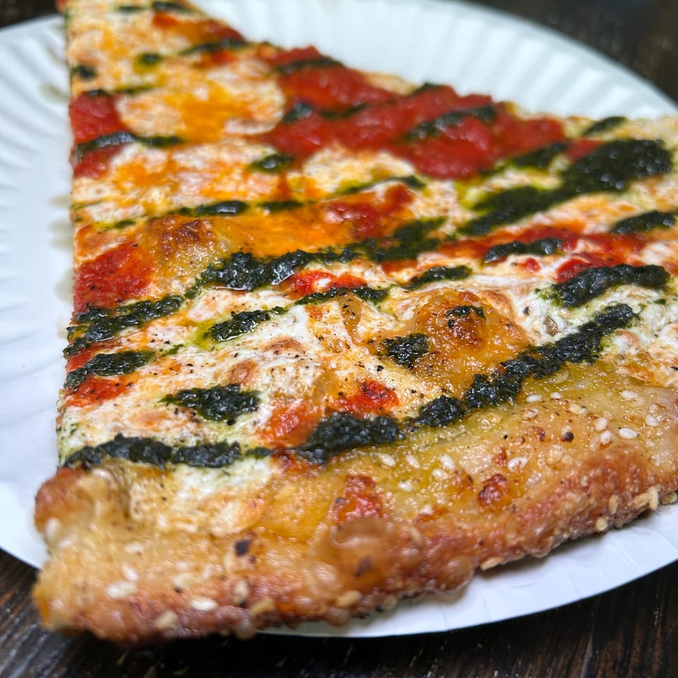 A slice of The Sauce Pizzeria's pizza with red sauce, melted cheese, and green pesto stripes.