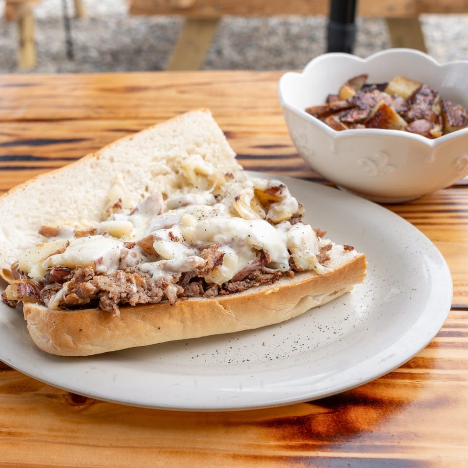 Cheesesteak sandwich with melted cheese and onions, served with a side of roasted potatoes.